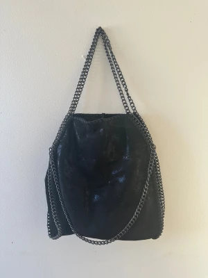 Svart axelväska med kedjedetaljer - Cool svart axelväska med glansig finish och grova kedjor som handtag. Väskan har en mjuk, mockaliknande yta och snörning upptill för att stänga. Perfekt storlek för att få plats med allt du behöver och ger en edgy vibe till din outfit.