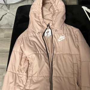 Ljusrosa pufferjacka från Nike XS - Säljer en ljusrosa pufferjacka från Nike i storlek XS. Jackan har huva, dragkedja och Nike-logga på bröstet. Den är quiltad och har Therma-FIT-teknologi som håller dig varm. Perfekt för kyliga dagar och riktigt snygg streetstil.