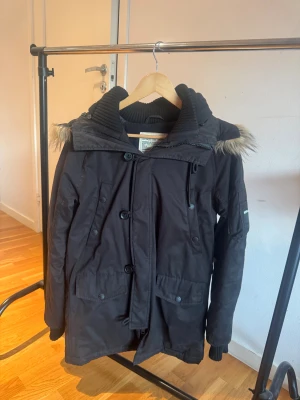 Svart vinterjacka från Replay - Snygg svart parkajacka från Replay med huva och fuskpälsdetalj. Jackan har flera fickor, knappar och dragkedja framtill samt ribbade muddar vid ärmslut. Perfekt för kalla dagar med sin varma och stilrena design.