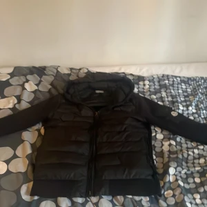 Svart pufferjacka från Moncler - Svart pufferjacka från Moncler med huva och quiltad front. Jackan har långa ärmar med Moncler-logga på fickan, dragkedja framtill och ribbade muddar. Materialet är polyester och dun, vilket gör den både lätt och varm. Perfekt för dig som vill ha en clean och stilren look. Där vid dragkedjan så finns de ingen sån man kan stänga fast de går frf att stänga och de inget man märker eller ser 