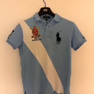 Blå Polo Ralph Lauren piké  - Snygg ljusblå pikétröja från Polo Ralph Lauren med vit diagonal rand och broderade detaljer på bröstet. Klassisk krage, korta ärmar och custom fit-passform. Ikonisk polospelare på bröstet och broderad siffra på ärmen. Perfekt för dig som gillar sportig stil.
