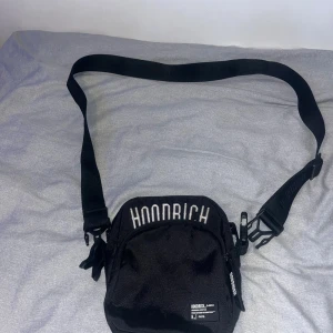 Svart axelväska från Hoodrich - Snygg svart axelväska från Hoodrich med justerbar rem och broderad logga i vitt framtill. Väskan har dragkedja och flera fack för dina prylar. Perfekt storlek för att bära med sig det viktigaste.
