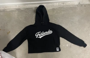 Svart Frölunda hoodie croppad dam S - Svart hoodie från Frölunda Indians i dammodell, storlek S. Croppad passform med stor vit Frölunda-logga på bröstet och emblem på nederkanten. Mjuk bomullskvalitet, huva med metallöljetter och långa ärmar. Perfekt för dig som vill visa support för Frölunda.