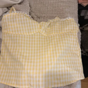 Gul och vit rutig linnetopp med spets - Säljer en söt linnetopp i gul och vit rutig design med tunna axelband och spetsdetaljer vid halsringningen. Toppen har en mjuk, feminin form och är perfekt för varma dagar. Passar dig som gillar ljusa färger och romantisk stil.