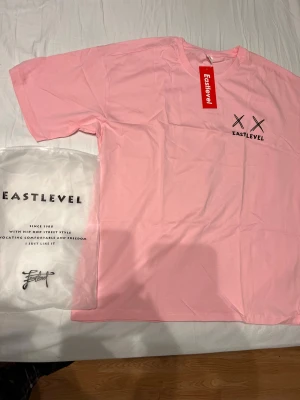 Rosa t-shirt från Eastlevel XL - Snygg rosa t-shirt från Eastlevel i storlek XL. Framsidan har tryck med två svarta kryss och logga, baksidan har ett stort smiley-tryck med korsade ögon. Skön passform och tillverkad i mjuk bomull. Perfekt för dig som gillar streetstyle och vill sticka ut.