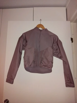 Gammaldagsrosa vindjacka Nike Running Division - Endast provad. Säljer en gammaldagsrosa vindjacka från Nike Running Division med hel dragkedja framtill och hög krage. Jackan har långärmad design och är tillverkad i lätt syntetmaterial, perfekt för löpning och utomhusaktiviteter. Snygg och sportig look med diskreta reflexdetaljer.