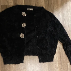 Svart fluffig kofta från Zara med blommiga knappar - Supermysig svart kofta från Zara i fluffigt pälsliknande material. Koftan har långa ärmar och tre stora blommiga silverknappar framtill som ger en lyxig vibe. Perfekt för dig som vill sticka ut med coola detaljer och mjuk känsla.
