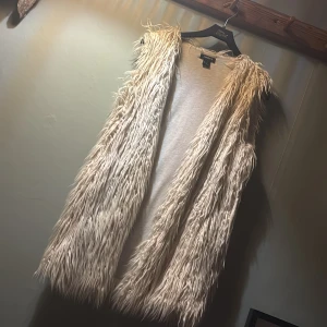 Beige lång pälsväst från Lindex - Säljer en beige lång pälsväst från Lindex i storlek M. Västen har ett coolt, fluffigt och långhårigt pälsmaterial som ger en riktigt statement-look. Den är ärmlös och har öppen front utan knäppning, perfekt att slänga över en hoodie eller t-shirt för extra edge.💕