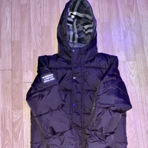 Svart pufferjacka från Burberry - Svart pufferjacka från Burberry med avtagbara ärmar så den kan användas som väst. Jackan har huva med klassiskt Burberry-mönstrat foder, snörning och dragkedja samt tryckknappar framtill. Patch med 'BURBERRY LONDON ENGLAND' på vänster ärm.storlek M men passar S men blir lite stor.en knapp är bort men man märker inte det är den högst upp.