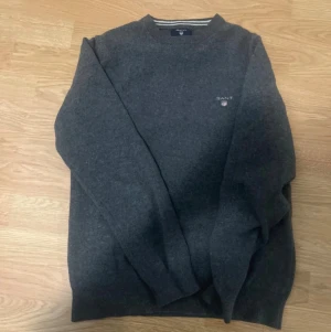 Mörkgrå stickad tröja från GANT - Mörkgrå stickad tröja från GANT i storlek S. Tröjan har rund hals med ribbad kant och vita ränder, samt GANT-logga broderad på bröstet. Perfekt för dig som gillar klassisk och enkel stil med snygga detaljer.