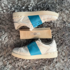 Valentino Open sneakers vit/blå, strl 43 - Snygga Valentino Open sneakers i vitt skinn med bred blå panel över vristen och ikoniska nitar på hälen. Klassisk rund tå, platt sula och vita snören. Perfekta för dig som vill sticka ut med en lyxig och modern look.