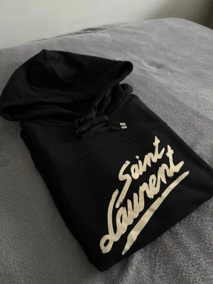 Saint Laurent tröja  - Svart hoodie från Saint Laurent med stor vit logotyp på bröstet. Tröjan har huva med snörning, magficka och långa ärmar. Tillverkad i mjuk bomull som är skön att ha på sig. Perfekt för dig som gillar streetwear och vill ha en snygg statement-tröja.