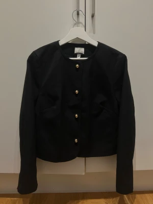Svart kavaj med guldknappar - Stilren svart kavaj med rund halsringning och fyra snygga guldknappar framtill. Plagget har lång ärm och en rak, något kortare passform som ger en clean look. Perfekt för dig som gillar klassisk men trendig stil.
