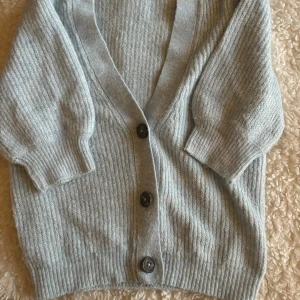 Ljusgrå kofta från JDY med knappar - Mysig ljusgrå kofta från JDY i stickad kvalitet. Koftan har ribbade muddar, v-ringad hals och tre stora svarta knappar framtill. Perfekt att slänga över en t-shirt för en chill look. Passar dig som gillar enkel och stilren design.