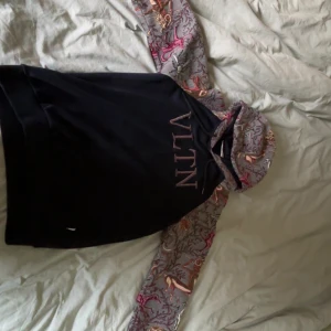 Valentino hoodie med mönstrade ärmar - Unik hoodie från Valentino med svart kropp och gråmelerade ärmar och huva med broderat mönster i rött, orange och beige. Stor VLTN-logga framtill. Hoodien har långärmade raglanärmar och huva med snyggt mönster som sticker ut. Perfekt för dig som gillar exklusiv streetwear.