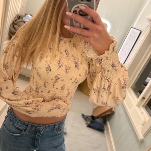 Beige blommig blus med puffärm - Supersöt beige blus med blommigt mönster och croppad passform. Blusen har långa puffärmar med smock vid ärmslut och över bröstet, vilket ger en snygg och feminin siluett. Perfekt att styla med jeans för en chill och trendig look.