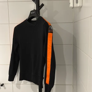 Svart fleece tröja Parajumpers - Svart långärmad fleece tröja från Parajumpers med orange detaljer längs axlar och ena ärmen. Snyggt tryck med Parajumpers-logga på axeln och text längs ärmen. Mjuk och skön passform, perfekt för dig som gillar sportig stil. Väldigt Fint skick size 140-146 nypris 1599, mitt pris 450!