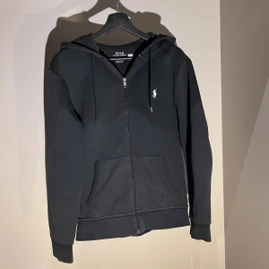  Polo Ralph Lauren zip hoodie - Svart hoodie från Polo Ralph Lauren med dragkedja och huva. snörning vid huvan och en vit broderad logga på bröstet. Praktisk känguruficka framtill och ribbade muddar. Perfekt för chill dagar. Hör av dig via dm vid frågor och funderingar🤗