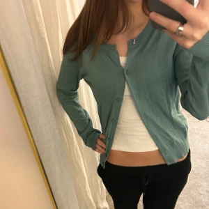 Grön kofta med knappar - Snygg grön kofta med rund hals och långa ärmar. Koftan har en clean look med knappar hela vägen framtill och ribbade muddar vid ärmslut och nederkant. Perfekt att slänga över en topp för en chill och stilren vibe.