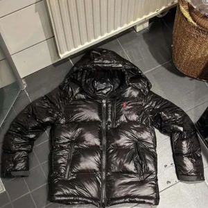 Svart pufferjacka från Polo Ralph Lauren - Säljer en svart pufferjacka från Polo Ralph Lauren med huva och glansig finish. Jackan har dragkedja framtill, flera fickor med dragkedja och den klassiska röda Polo-loggan på bröstet. Perfekt för kalla dagar och riktigt snygg streetstil.