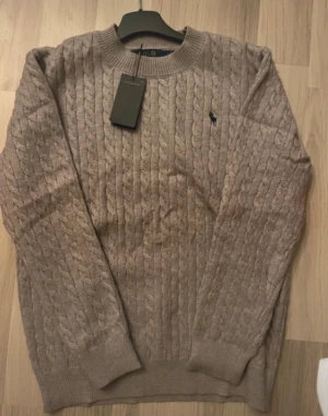 Grå kabelstickad tröja Polo Ralph Lauren - Snygg beige kabelstickad tröja från Polo Ralph Lauren med klassisk rund hals och broderad logga på bröstet. Tröjan har ribbade muddar vid ärmslut och nederkant, perfekt för dig som gillar stilrena och tidlösa plagg.