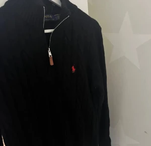 Svart stickad half zip tröja Polo Ralph Lauren - Svart kabelstickad tröja från Polo Ralph Lauren med röd broderad logga på bröstet. Tröjan har en ribbad hög krage och half zip-dragkedja med läderdetalj. Perfekt för dig som gillar klassisk stil med en modern touch.