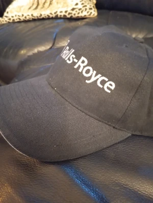 Svart Rolls-Royce keps - Snygg svart keps med vit Rolls-Royce-logga broderad framtill. Klassisk böjd skärm med vit kant och justerbar passform. Perfekt för dig som gillar exklusiva bilar och vill ha en stilren accessoar. Materialet känns som bomull och kepsen har en tidlös look.