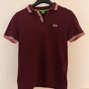 Vinröd kortärmad skjorta från Hugo Boss - Vinröd skjorta från Hugo Boss med korta ärmar och vit broderad logga på bröstet. Kragen och ärmsluten har snygga ränder i vitt och rött som ger en cool kontrast. Skjortan har knappar vid halsen och är tillverkad i mjuk bomull för skön känsla.