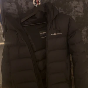 Svart dunjacka från Sail Racing - Svart dunjacka från Sail Racing med 700 FP dunfyllning. Jackan har huva, dragkedja och logga på bröstet. Perfekt för kalla dagar och har en clean, sportig look. Materialet är polyester och dun, och modellen är normal i passningen.