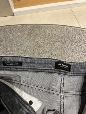 Grå tapered jeans från Jack & Jones - Snygga grå jeans från Jack & Jones, modell Tapered/Mike. De har en modern tapered passform med smalare ben och klassisk femficksdesign. Jeansen är tillverkade i mjukt denim och har subtila slitningar för en cool look. 32 - midja.     30- innerbenslängd dvs 32/30