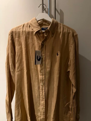 Linneskjorta från Ralph Lauren - Snygg beige linneskjorta från Ralph Lauren. Skjortan har aldrig använts, lappen sitter kvar som syns på bilderna. Vid frågor är det bara att höra av sig!
