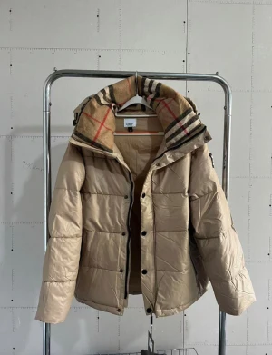 Beige pufferjacka från Burberry - Snygg beige pufferjacka från Burberry med avtagbara ärmar så den kan bäras som väst. Jackan har klassiskt rutigt foder i huvan, svarta dragkedjor och tryckknappar samt en tydlig Burberry-patch på bröstet. Perfekt för kalla dagar med sin vadderade design.