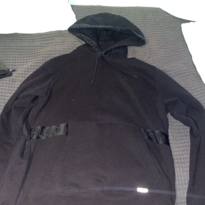 Svart hoodie från Hugo Boss - Svart hoodie från Hugo Boss med snygga ton-i-ton detaljer och logoband över magen. Tröjan har huva med snörning, lång ärm och en klassisk känguruficka framtill. Perfekt för dig som gillar stilrena och bekväma plagg.