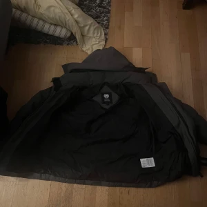 Svart dunjacka från Canada Goose - Svart dunjacka från Canada Goose med klassisk patch på ärmen. Jackan har huva, dragkedja och knappar framtill. Fodrad och rejält puffad för att hålla värmen under kalla dagar. Perfekt för dig som vill ha en stilren och funktionell vinterjacka.