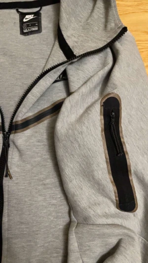 Grå hoodie från Nike med dragkedja - Grå hoodie från Nike i storlek S med hel dragkedja framtill och svart Nike-logga på bröstet. Tröjan har huva, långärmade ärmar och en cool svart detalj med dragkedja på ena ärmen. Svarta och bruna linjer går över bröstet och ärmen för extra stil.