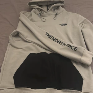 Beige hoodie från The North Face - Säljer en beige hoodie från The North Face med svart magficka och logga på bröstet samt tryck på ärmen. Tröjan har huva med snörning och ribbade muddar. Perfekt för chill dagar och streetwear-stil.