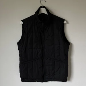 Svart quiltad dunväst från J.Lindeberg - Svart dunväst från J.Lindeberg med ståkrage och dragkedja framtill. Quiltad design med två snedställda fickor och diskret logotyp på insidan. Tillverkad i 100% polyester med Primaloft-fyllning för extra värme. Perfekt att slänga över en hoodie när det är kyligt ute. Modell: Gavin Compact Melton - 5I14