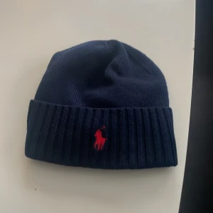 Mörkblå ullmössa från Polo Ralph Lauren - Snygg mörkblå mössa från Polo Ralph Lauren i 100% ull. Klassisk ribbad kant och ikonisk röd Polo-logga broderad framtill. Perfekt för dig som vill ha en stilren och varm mössa med premiumkänsla och diskret branding. Mössan är nästan helt oanvänd och köpt på kidsbrandstore.Storlek ca 14 åringar passar äldre och yngre också. Ny pris 700.