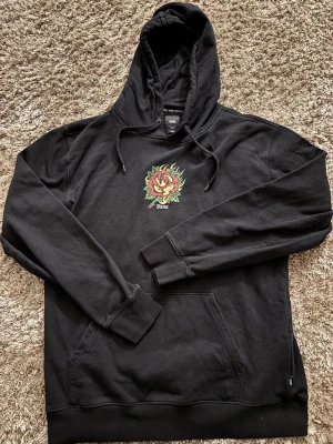 Svart Vans hoodie med rosprint - Svart hoodie från Vans i storlek L med en fet rosprint i rött, gult och grönt på bröstet. Klassisk huva med snörning och stor magficka. Mjuk insida och ribbade muddar. Perfekt för dig som gillar streetwear och coola detaljer. Tröjan är i ett väldigt bra skick. Har du några frågor så är det bara att skriva.
