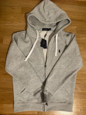 Grå hoodie från Polo Ralph Lauren - Snygg grå hoodie från Polo Ralph Lauren med klassisk broderad logga på bröstet. Tröjan har huva med vita snören, hel dragkedja och två fickor framtill. Mjuk bomullsmix som är perfekt för chill eller häng. Stilsäker och enkel att matcha med jeans eller joggers.