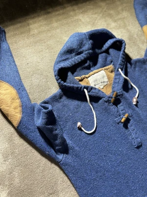 Blå hoodie med snörning från Angelo Litrico - Snygg blå hoodie från Angelo Litrico med beige armbågslappar och vita snören med trädetaljer. Tröjan är stickad och har en stor känguruficka framtill. Perfekt för dig som gillar chill och avslappnad stil.