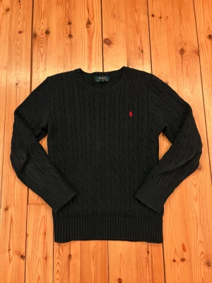 Svart stickad tröja Polo Ralph Lauren - Marinblå stickad tröja från Polo Ralph Lauren med klassiskt kabelstickat mönster och rund hals. Tröjan har långa ärmar och ribbade muddar vid ärmslut och nederkant. På bröstet finns den ikoniska röda Polo-loggan broderad. Perfekt för dig som gillar stilren och tidlös design.        Strorlek: 14-16år.                                                                   Tröjan är marinblå men ljuset på bilder får den att se mörkare ut.