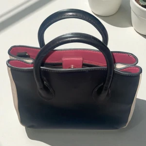 Svart/beige handväska i skinn från Zara - Snygg handväska i svart och beige skinn med röd insida, från Zara Basic Collection. Väskan har två handtag, magnetlås, en stor innerficka med dragkedja, samt tre andra fickor. Stilren design med kontrastfärger och metallfötter undertill. Lite repig yta på vissa ställen men inget som syns på håll!