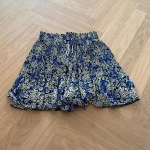 Blommiga plisserade shorts från FBSSTER - Säljer ett par blommiga shorts från FBSSTER i storlek XXS. De har hög midja med smock, plisserad utsida och svart innershorts. Färgerna går i blått, lila, grönt och vitt mot svart botten. Perfekta för dig som gillar färg och mönster!