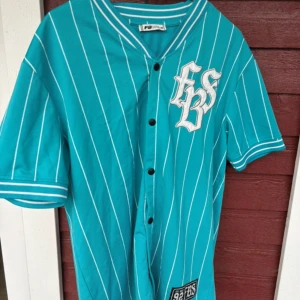 Turkos randig baseballskjorta FB Sister - Turkos baseballinspirerad skjorta från FB Sister i storlek XS. Skjortan har vita ränder, korta ärmar och knappar framtill. Broderad logga på bröstet och patch nertill. Ribbad krage och ärmslut ger en sportig vibe. Perfekt för dig som gillar streetstyle.(OVERSIZED)!!