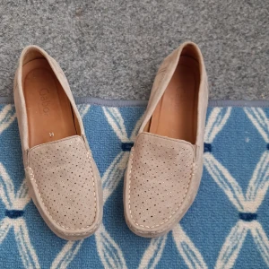 Beige mockaloafers från Gabor - Snygga beige loafers från Gabor i mjuk mocka med små silvriga nitar på ovandelen. Skorna har platt sula, rund tå och klassisk söm runt tån. Perfekta för dig som gillar stilrena och bekväma skor med lite extra detalj.