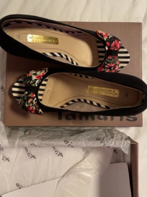 Svarta pumps med blommig rosett Tamaris - Unika pumps från Tamaris i svart mocka med öppen tå och hög klack. Skorna har en snygg rosett framtill i blommigt tyg med inslag av rött, grönt och vitt samt randiga detaljer. Insidan har också ett randigt mönster som ger extra edge.