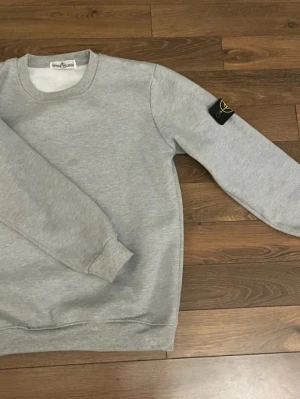 Grå sweatshirt från Stone Island - Snygg grå sweatshirt från Stone Island med klassisk rund hals och ribbade muddar. Tröjan har den ikoniska svarta patchen med gul kompasslogga på vänster ärm. Perfekt för en stilren och avslappnad look.