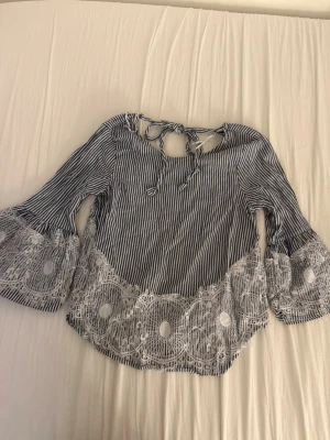 Randig blus med spetsdetaljer - Snygg blus med smala blå och vita ränder, vida trumpetärmar och dekorativa vita spetsdetaljer längs nederkant och ärmslut. Blusen har knytband i nacken och lätt böljande passform. Perfekt för en trendig och avslappnad look. Har en liten sminkfläck se bild❤️