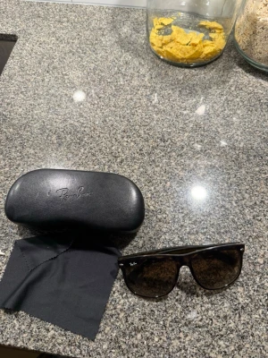 Ray-Ban solglasögon med fodral - Snygga Ray-Ban solglasögon säljes med original svart hårt fodral och putsduk. Perfekt accessoar för att lyfta din stil. Klassisk design och hög kvalitet från ett av de mest ikoniska märkena inom solglasögon.